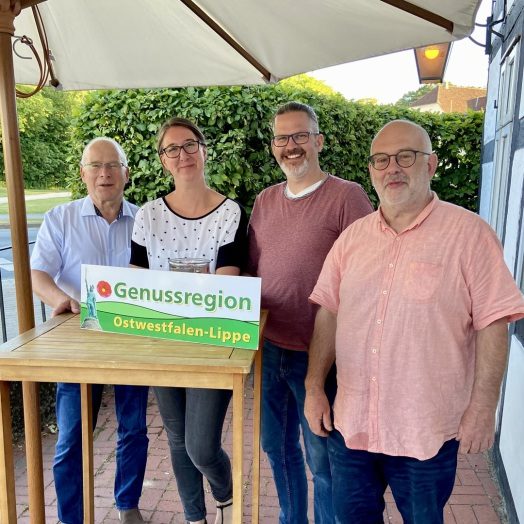 3 Männer und 1 Frau an einem Stehtisch stehend. Auf dem Stehtisch das Schild Genussregion Ostwestfalen-Lippe
